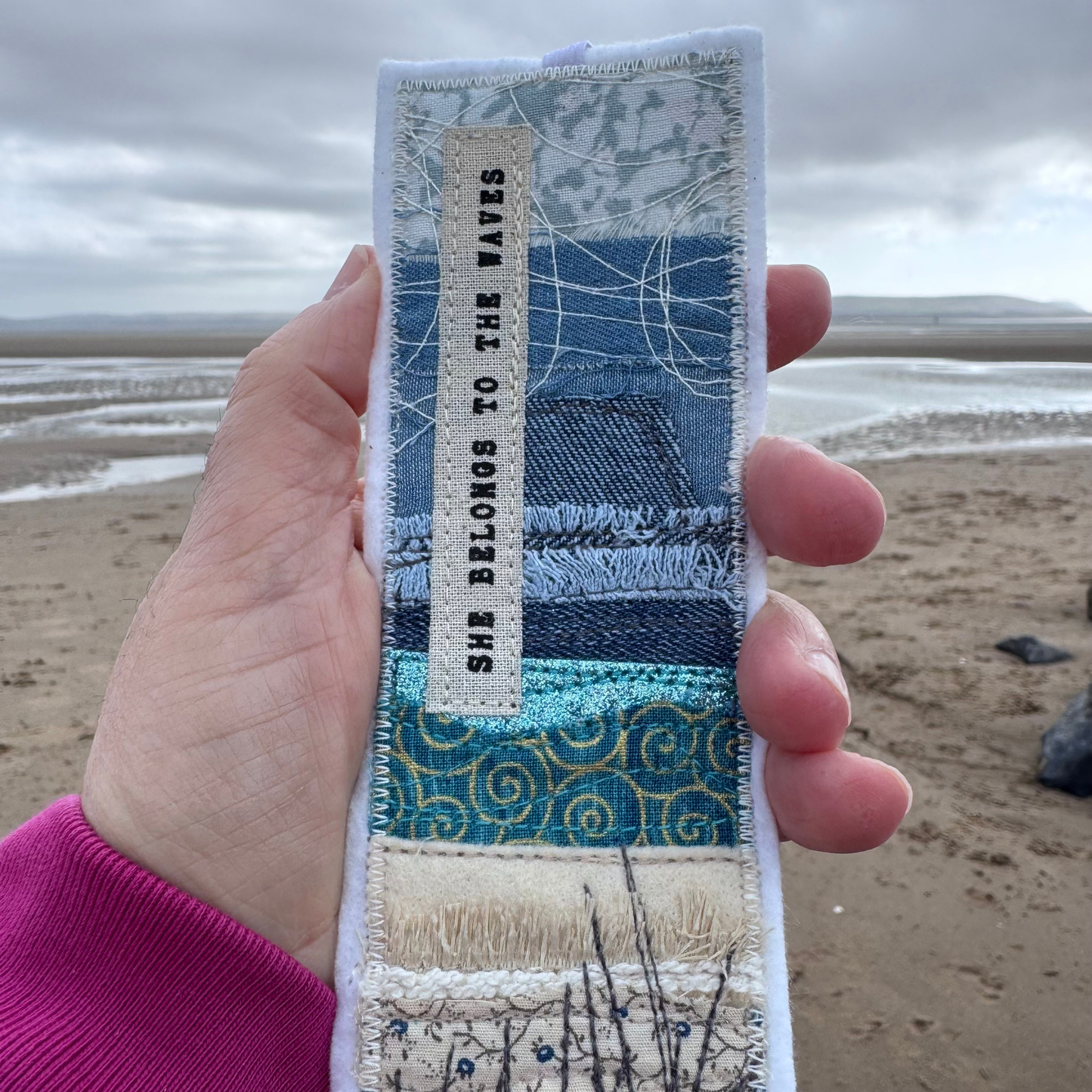 Close-up of a stitched fabric bookmark with elastic, held by the sea, featuring textured shoreline layers and a printed quote — a handmade wild water keepsake gift for book lovers.