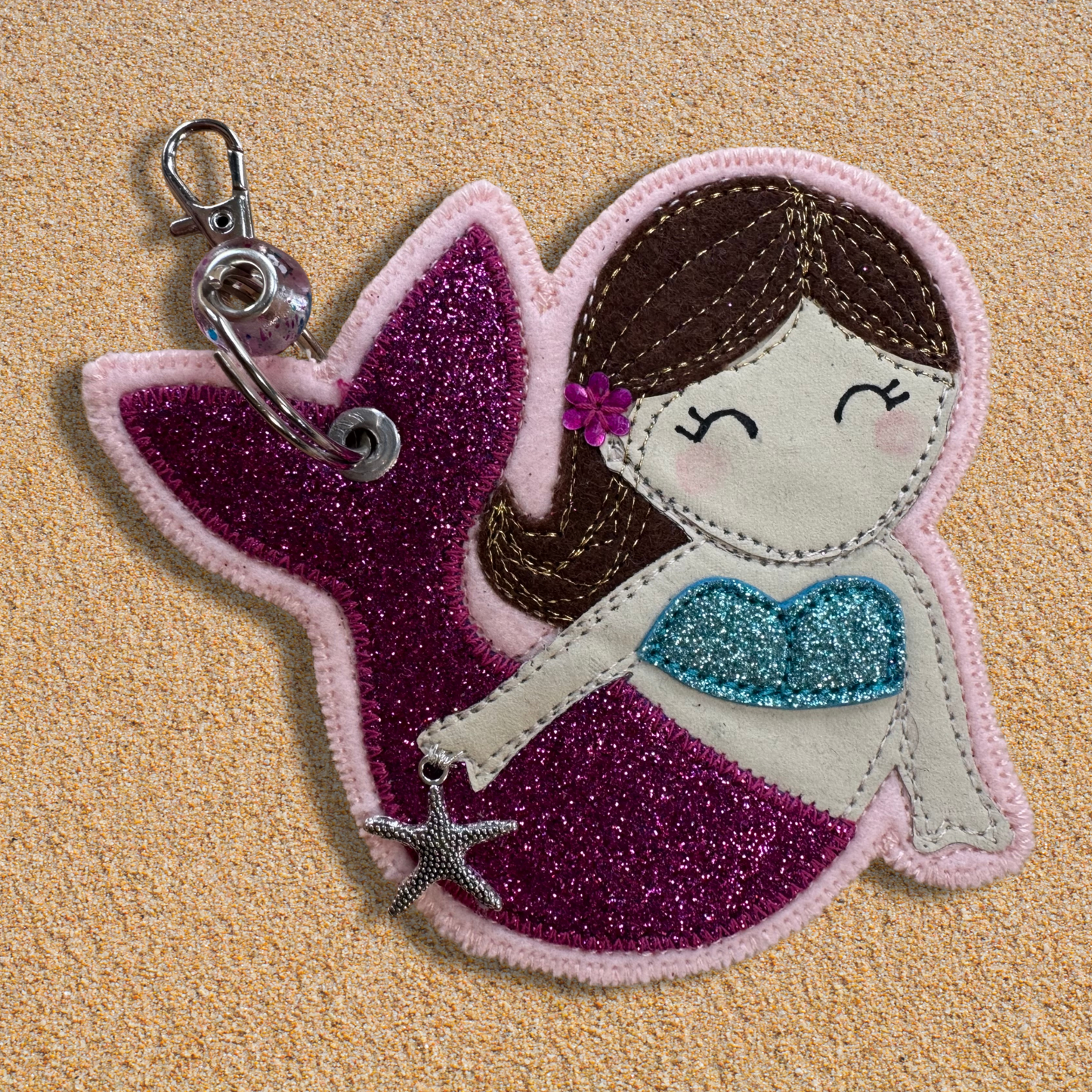 Handmade fabric mermaid charm named Meryl the Mermaid resting on soft golden sand, featuring a glittery pink tail, blue top, and brown hair with flower detail. Key chain clasp has a Murano glass bead. Perfect for hanging from your wild swimming kit bag or changing robe.