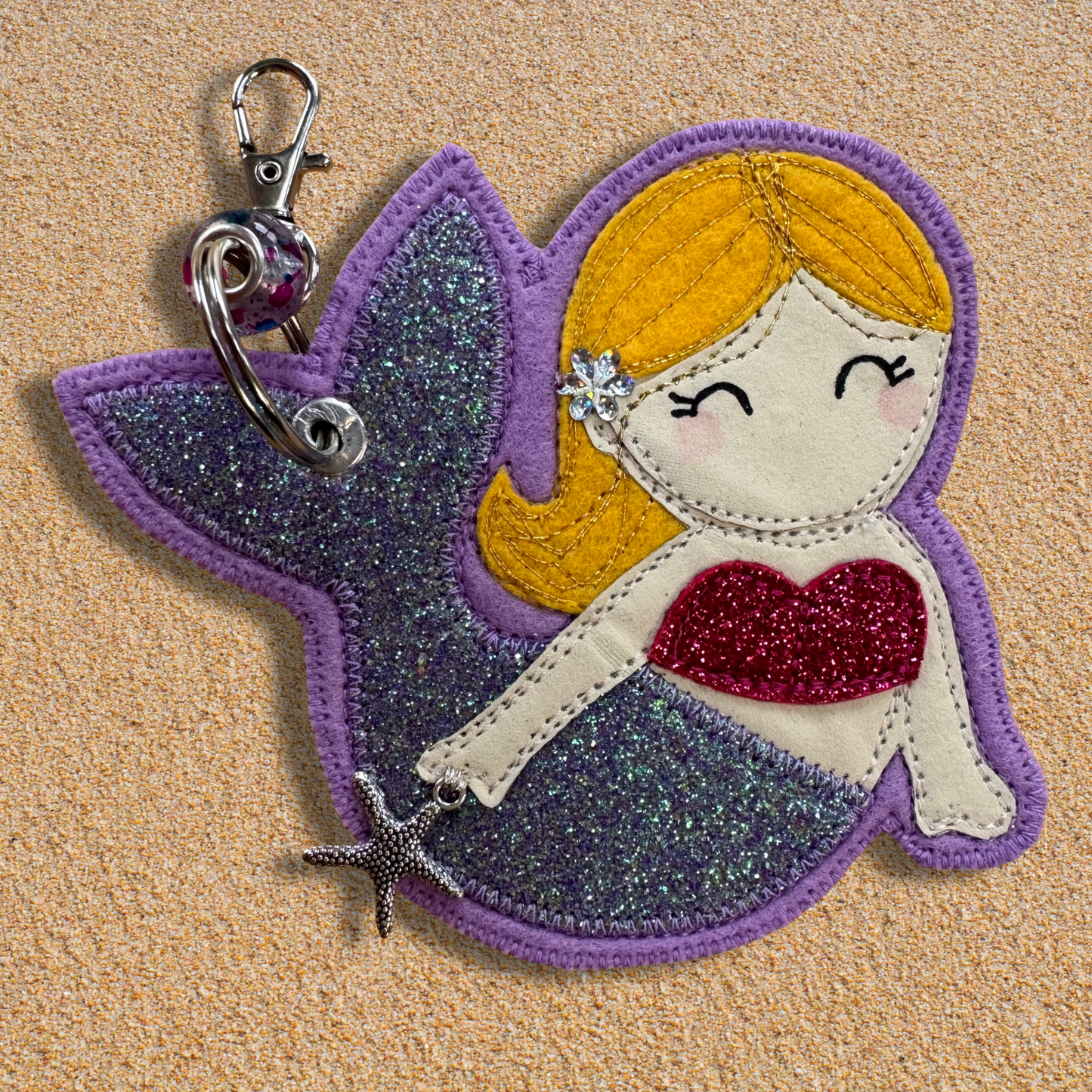 Handmade fabric mermaid charm named Meryl the Mermaid resting on soft golden sand, featuring a glittery lilac tail, pink top, and yellow hair with flower detail. Key chain clasp has a Murano glass bead. Perfect for hanging from your wild swimming kit bag or changing robe.