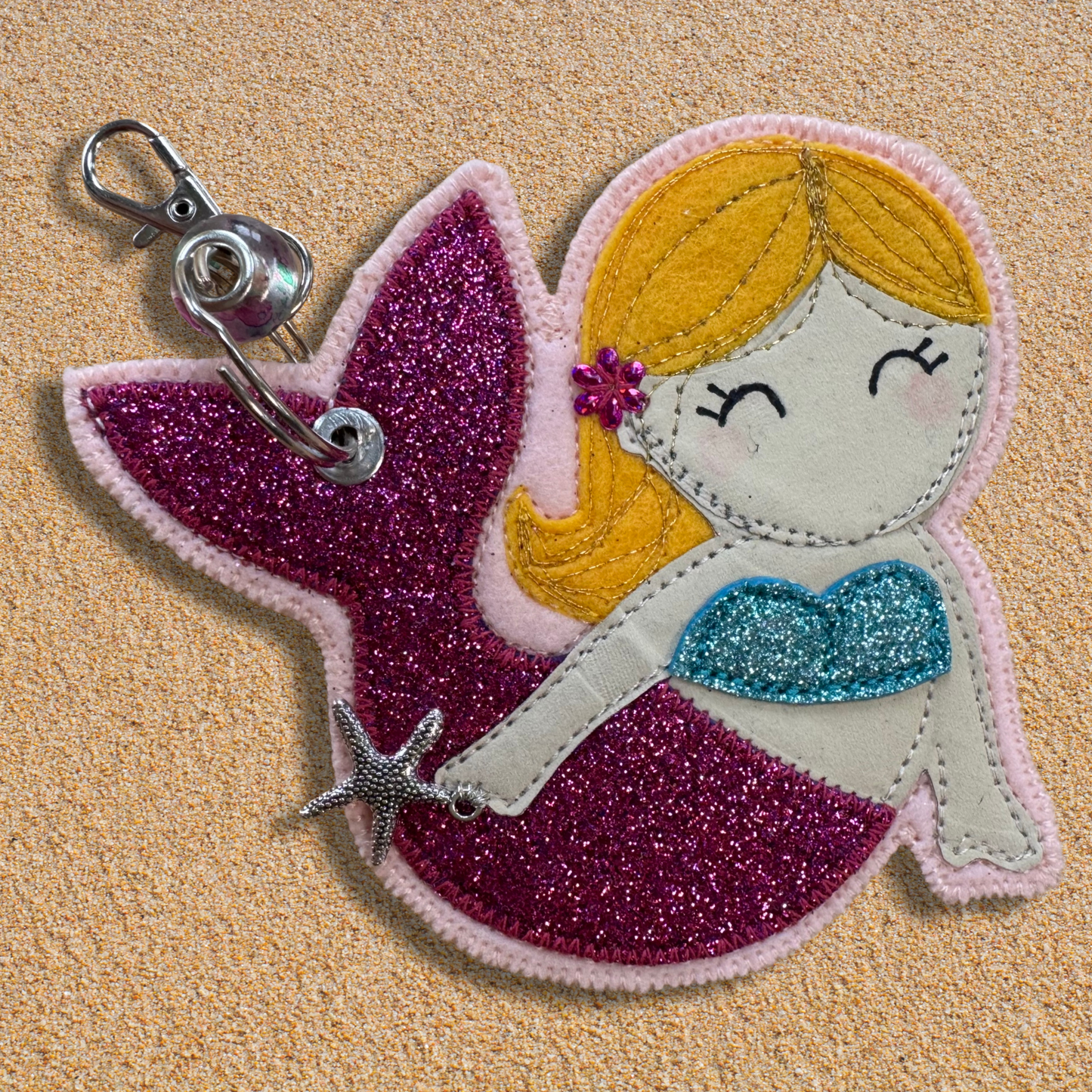 Handmade fabric mermaid charm named Meryl the Mermaid resting on soft golden sand, featuring a glittery pink tail, blue top, and yellow hair with flower detail. Key chain clasp has a Murano glass bead. Perfect for hanging from your wild swimming kit bag or changing robe.