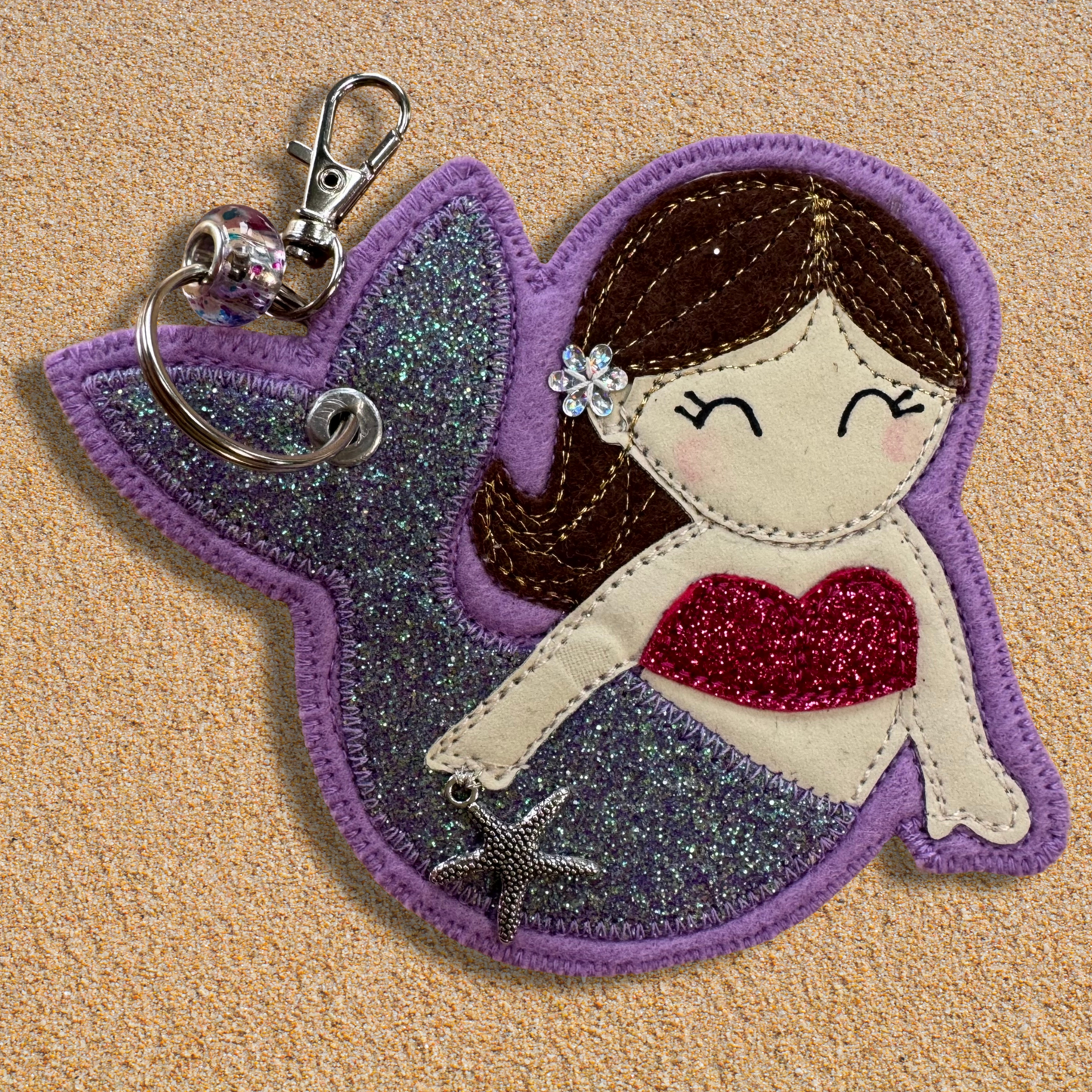 Handmade fabric mermaid charm named Meryl the Mermaid resting on soft golden sand, featuring a glittery lilac tail, pink top, and brown hair with flower detail. Key chain clasp has a Murano glass bead. Perfect for hanging from your wild swimming kit bag or changing robe.