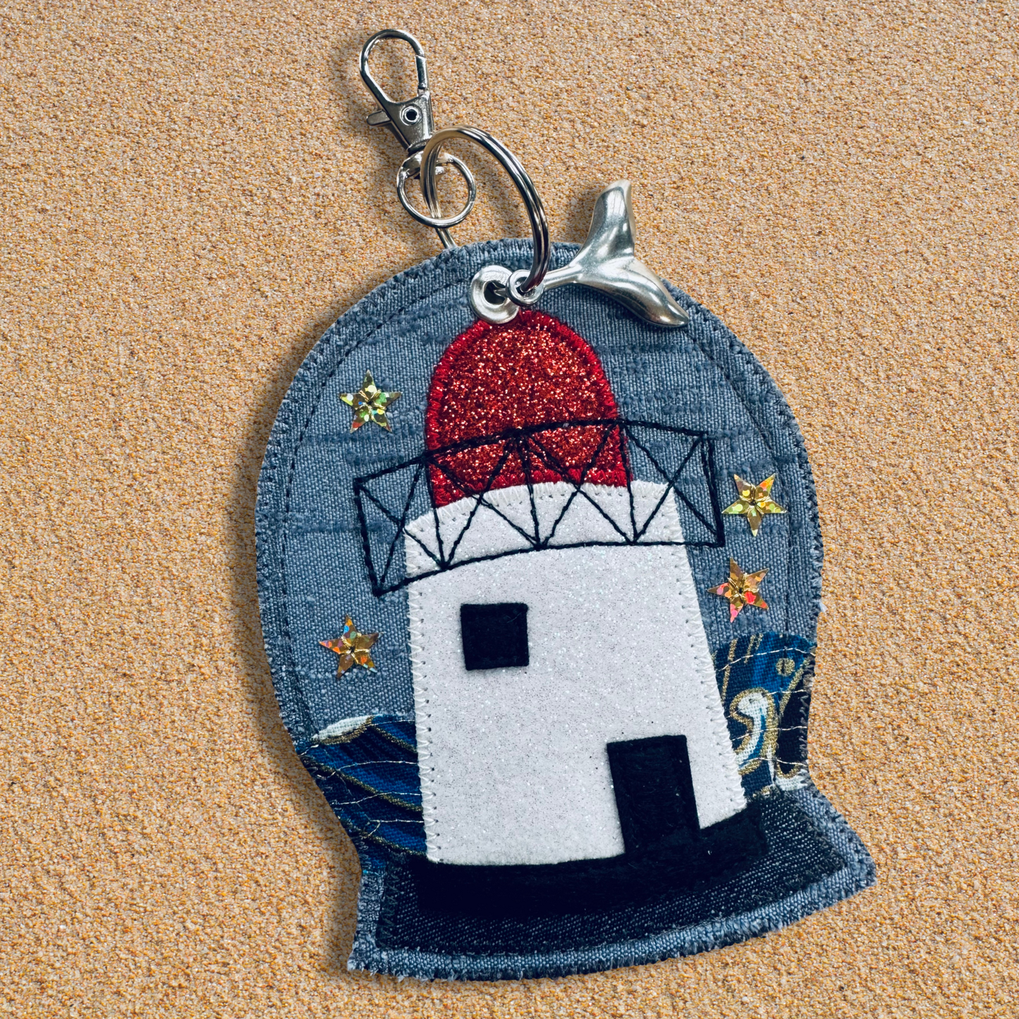 Close-up of the Waves & Wild Water Burry Port Lighthouse keyring charm, showing its glitter dome, denim base, and silver whale-tail detail.