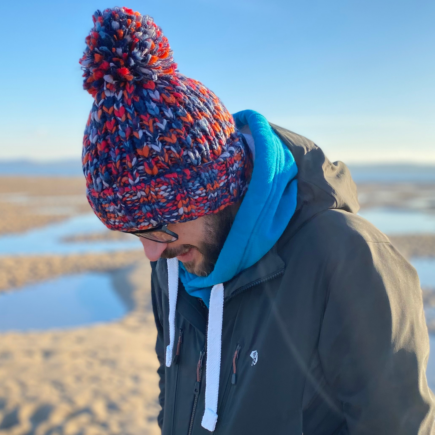 A man on the beach wearing a chunky beanie hat with a pom pom. 