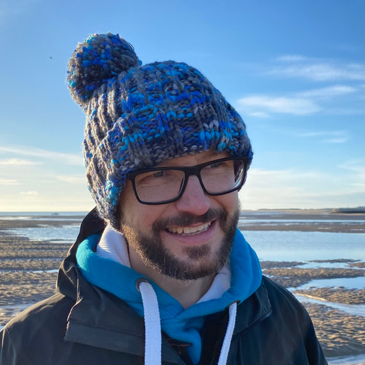 A man on the beach wearing a chunky knitted beanie hat with a pom pom.