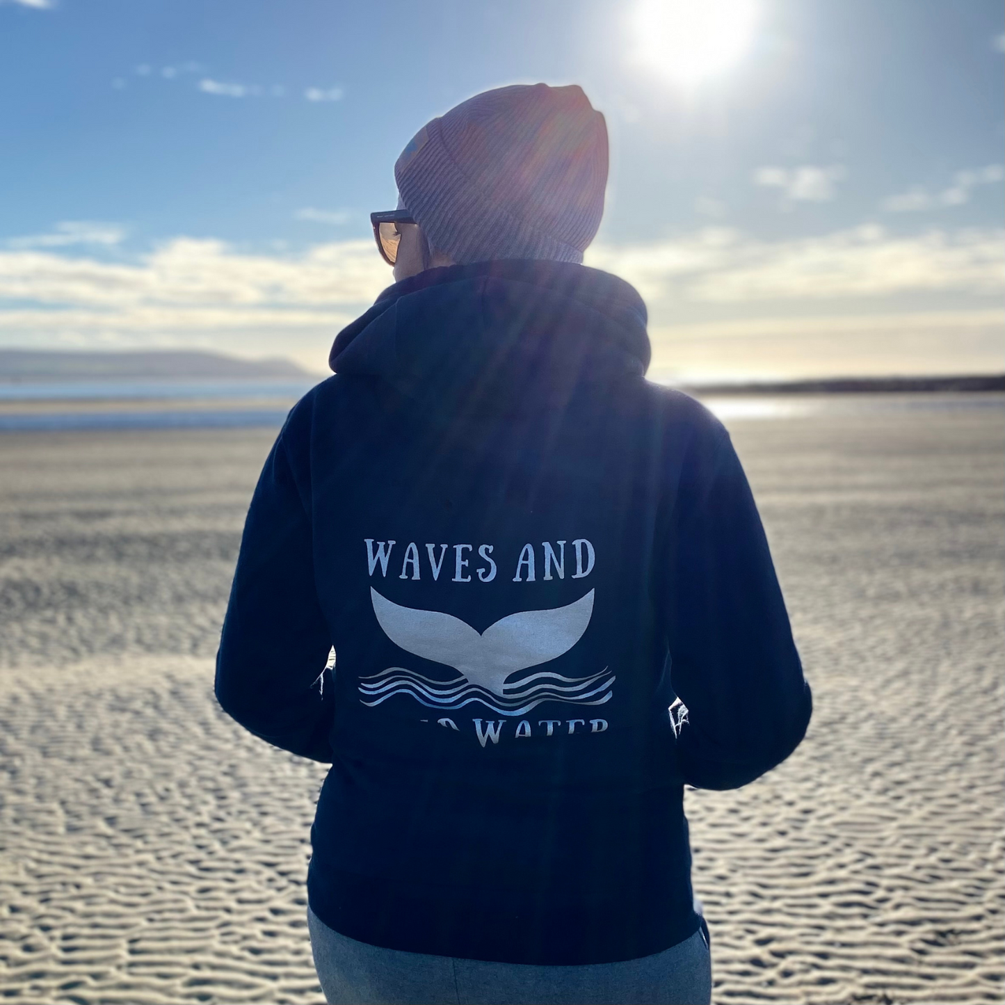 The back of a woman wearing a navy blue hoodie, with Waves & Wild Water logo printed on it in metallic silver ink.