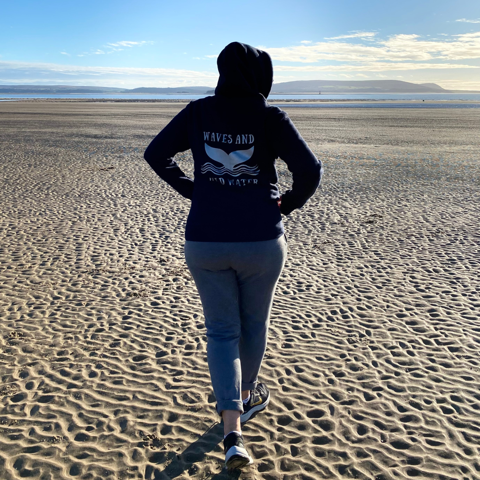 The back of a woman standing on a beach wearing a Waves & Wild Water hoodie.