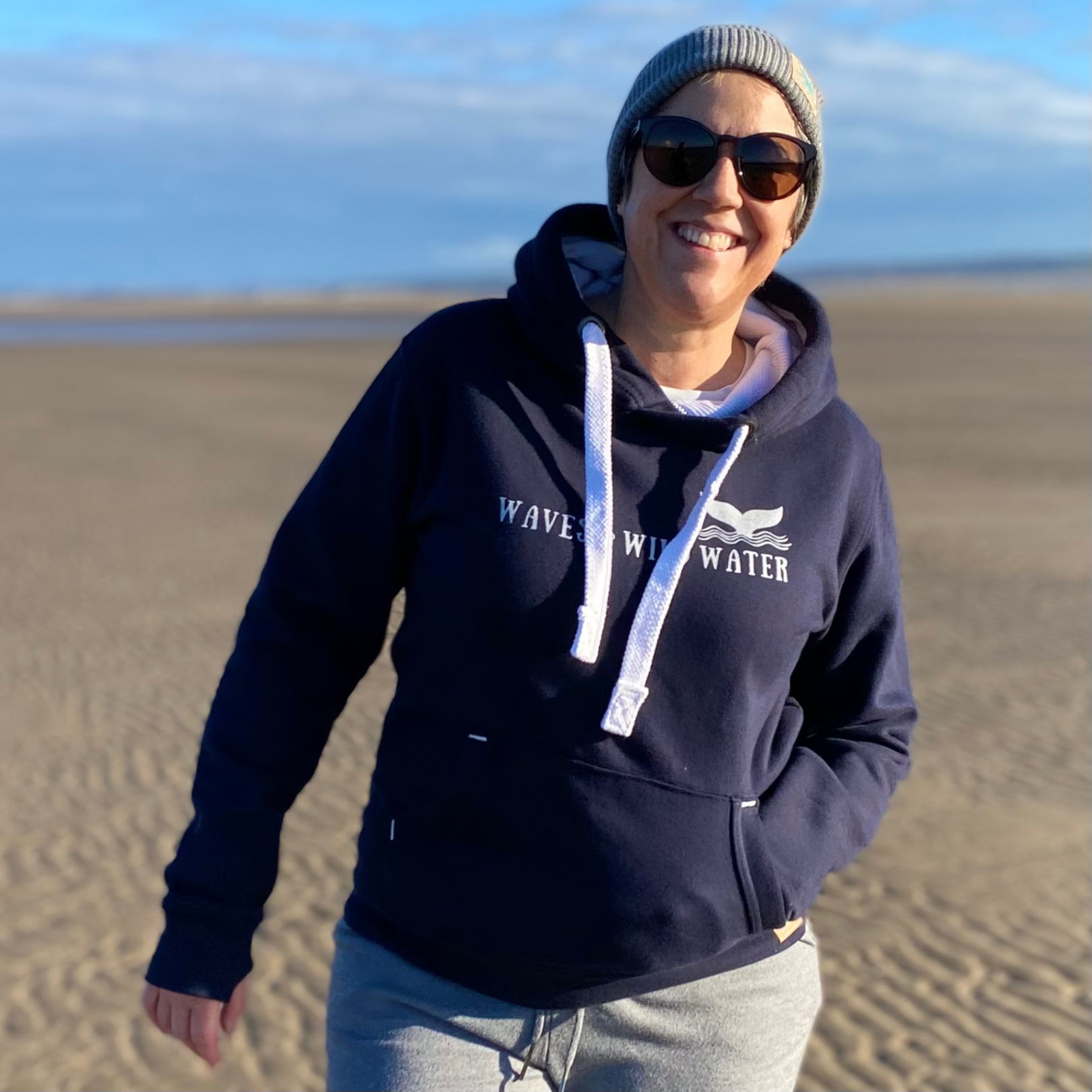A woman on a beach wearing a navy blue hoodie with Waves & Wild Water printed across the front in metallic silver ink.