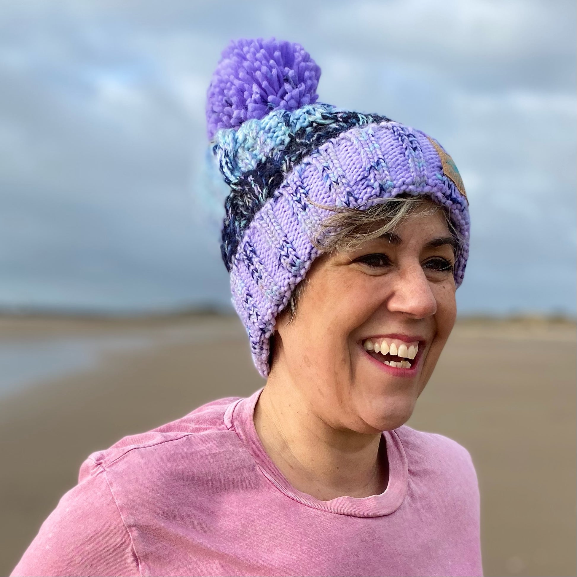 Woman wearing a lilac and blue knitted beanie hat with a pom pom.