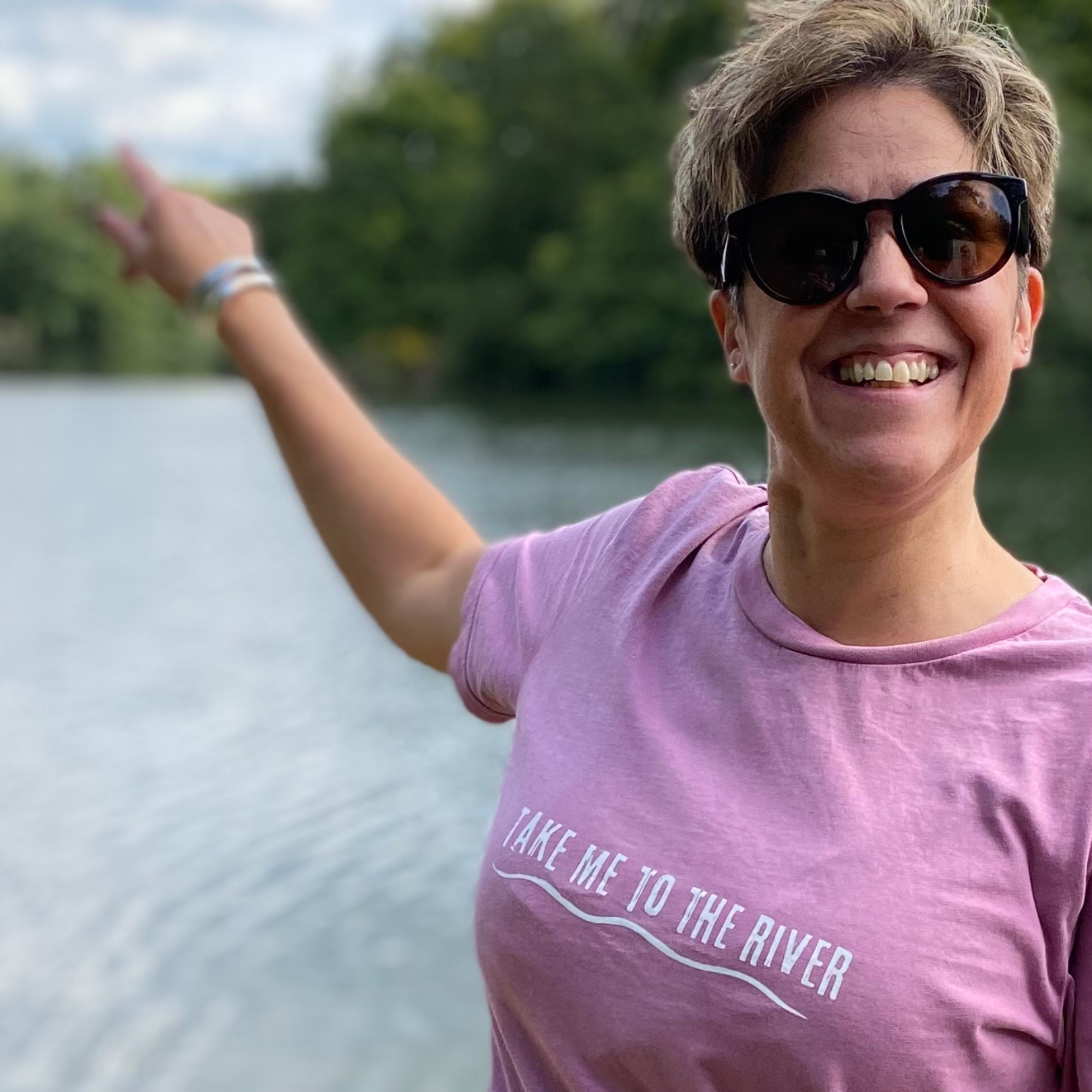 A woman wearing a pink t-shirt with 'Take me to the river' printed across the front while pointing at a river.