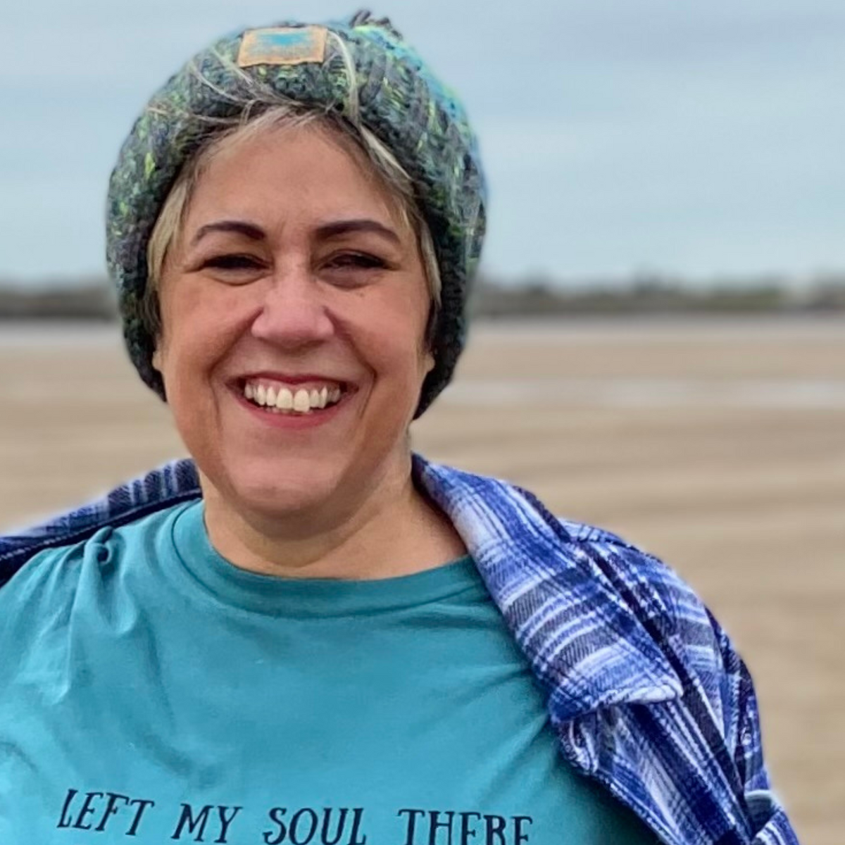 Wild swimmer Claire, shows off her stunning Lagoon knitted beanie hat during a windy walk on the beach. Knitted in shades of green and blue, it looks fab with our Waves and Wild Water range of t-shirts too.