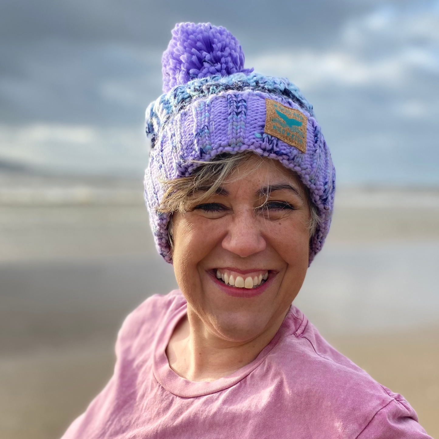 This is our gorgeous knitted beanie hat in lilac lagoon colours. It's called lilac lagoon because the chunky knit is in various shades of lilac and purple. It has a deep fold up cuff to keep your ears snug and cosy and is finished off with a plump woolly pom pom. Claire is wearing her hat on the beach after an invigorating swim in the sea.