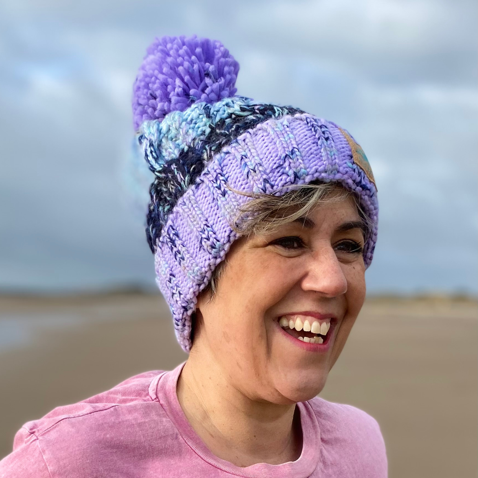 Wild swimmer Claire, shows off her stunning Lilac Lagoon knitted beanie hat during a windy walk on the beach. Knitted in shades of blue and lilac, it looks fab with our Waves and Wild Water range of t-shirts too.