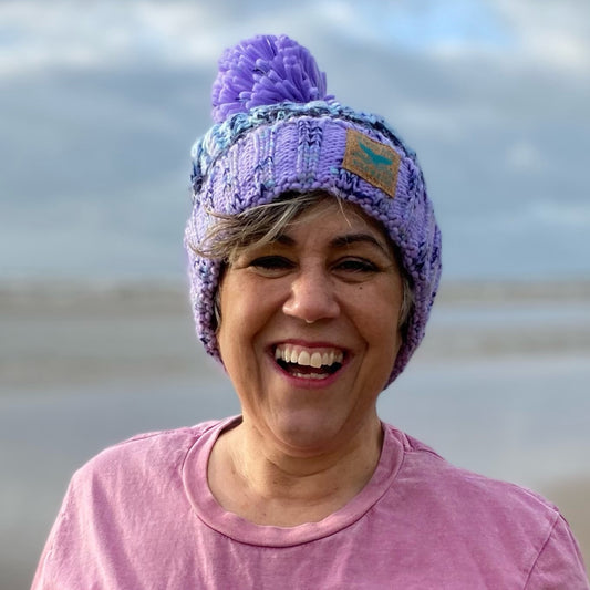 This is our gorgeous knitted beanie hat in lilac lagoon colours. It's called lilac lagoon because the chunky knit is in various shades of lilac and purple. It has a deep fold up cuff to keep your ears snug and cosy and is finished off with a plump woolly pom pom. Claire is wearing her hat on the beach after an invigorating swim in the sea.