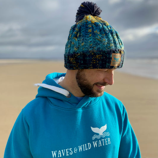 Glenn from Waves and Wild Water is wearing his knitted beanie hat in ocean blue. He's just been for a wild swim in the sea and is feeling cosy in his hat and his Waves and Wild Water hoodie in tropical waters blue.