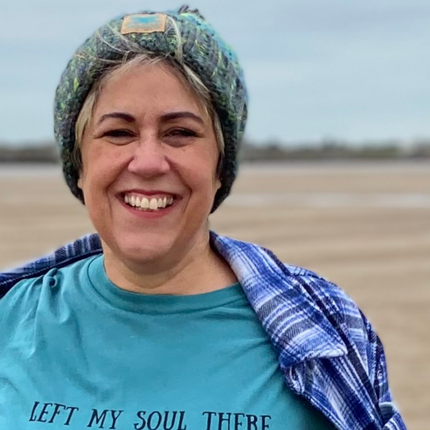 Wild swimmer Claire, shows off her stunning River Bed knitted beanie hat during a windy walk on the beach. Knitted in shades of green, blue and yellow, it looks fab with our Waves and Wild Water range of t-shirts too.