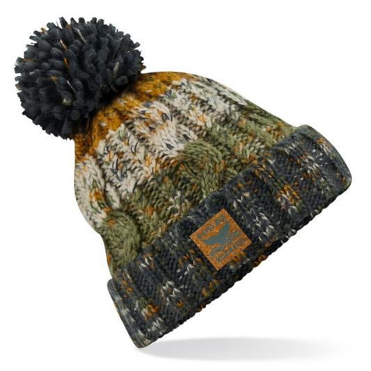 A marled knit beanie with a large dark navy pom-pom, featuring a pattern of olive green, mustard yellow, and cream, with a dark navy ribbed cuff and a small brown leather patch.