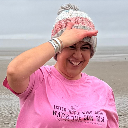 Waves and Wild Water owner Claire, tries to use her hand to shelter from the rain on the beach in Wales. She's wearing her Seashell Pink knitted beanie hat, and her pink Listen To The Wind Blow t-shirt. 