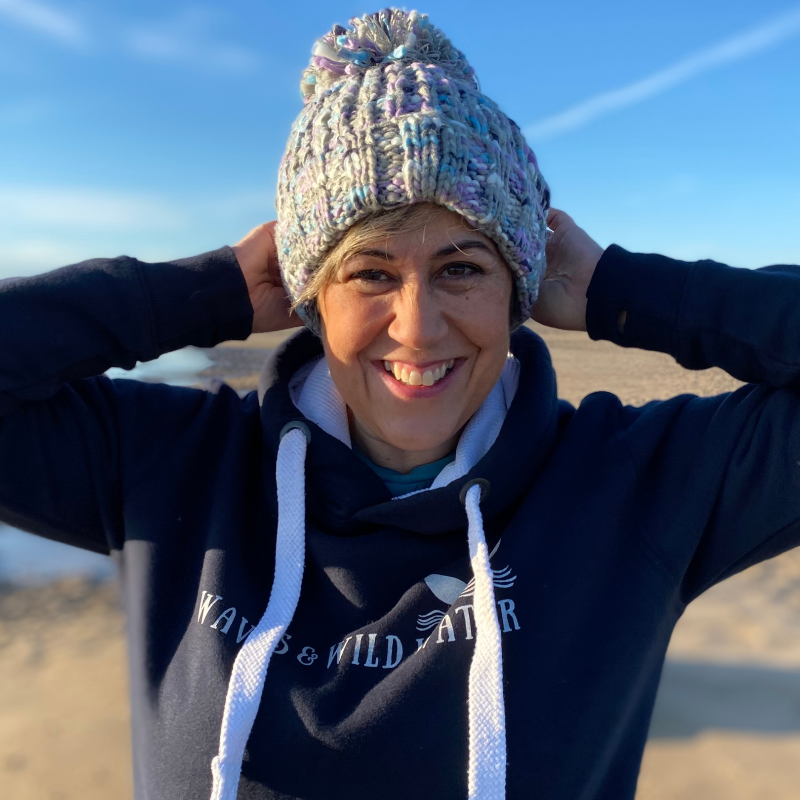 Wild swimmer and owner of Waves and Wild Water, Claire, enjoys a dose of winter on on the beach. She's cosy in her chunky knitted pom pom beanie hat, in shades of lilac and grey.