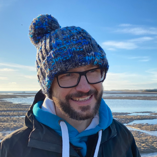 Waves and Wild Water owner Glenn, wears his chunky beanie hat with statement pom pom. Hand knitted in icy shades of blue and grey, this beanie is a favourite with wild swimmers, sea dippers, and cold water plungers.