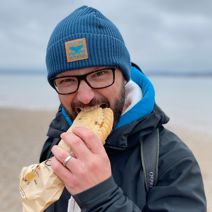 Waves and Wild Water owner Glenn, eats the obligatory post-swim hot pasty on the beach, whilst warming up in his Stormy Sea fisherman beanie.
