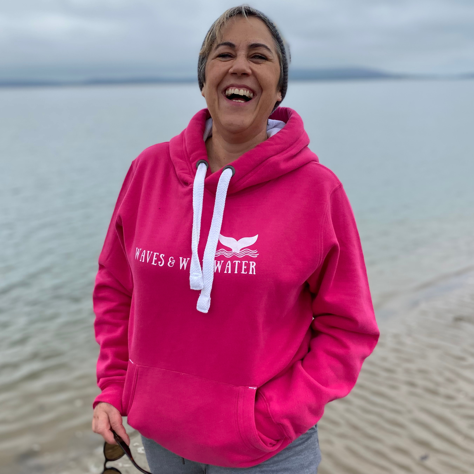 Wild swimmer and open water enthusiast, Claire, wears her sunset pink Waves and Wild Water hoodie on the beach. It's a grey day so she's happy she's wearing something so warm and snuggly.