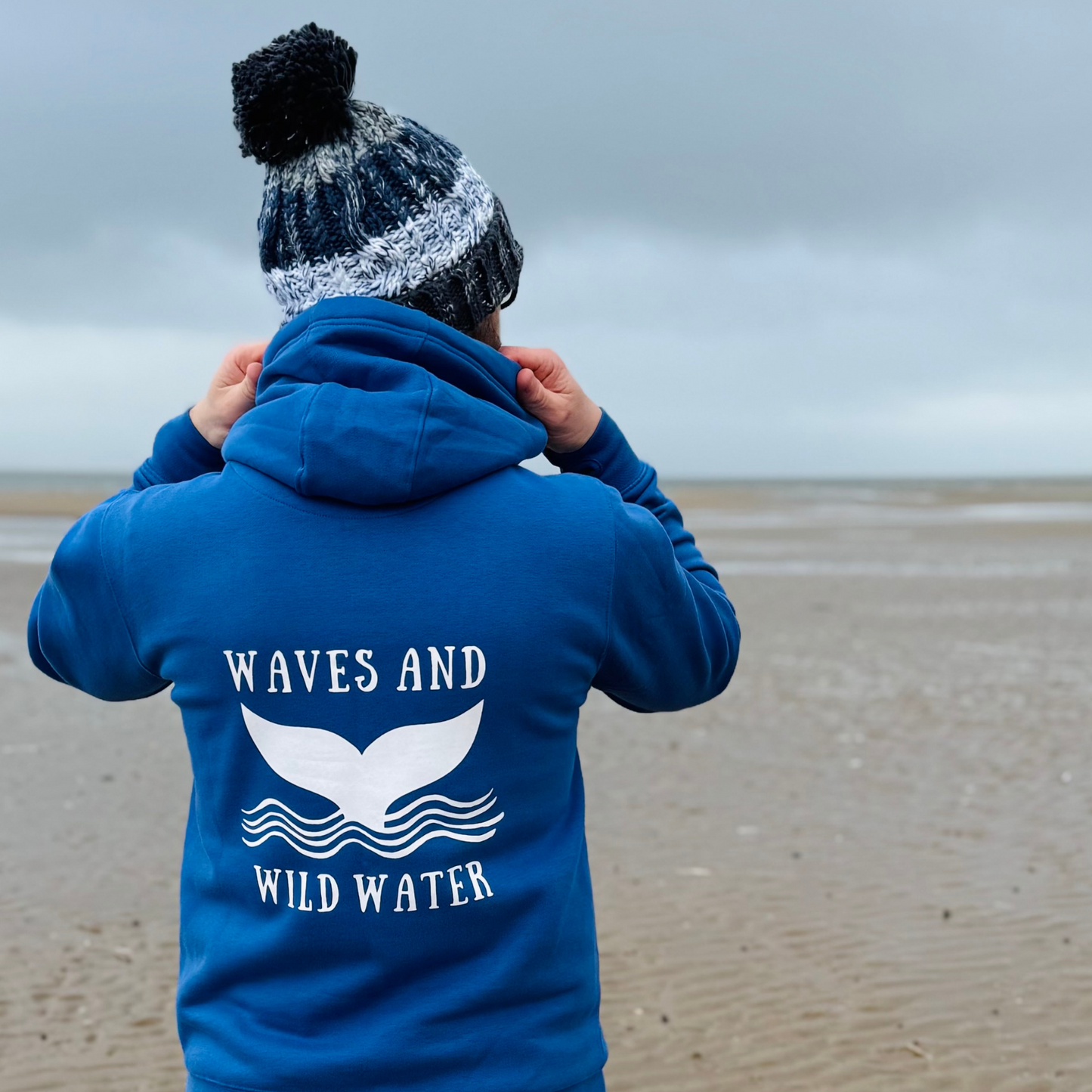 Waves and Wild Water owner and wild swimmer Glenn, shows off his stunning Swimmer Blue hoodie on the beach in South Wales. The bright white whale tail logo looks great against the bright blue hoodie.