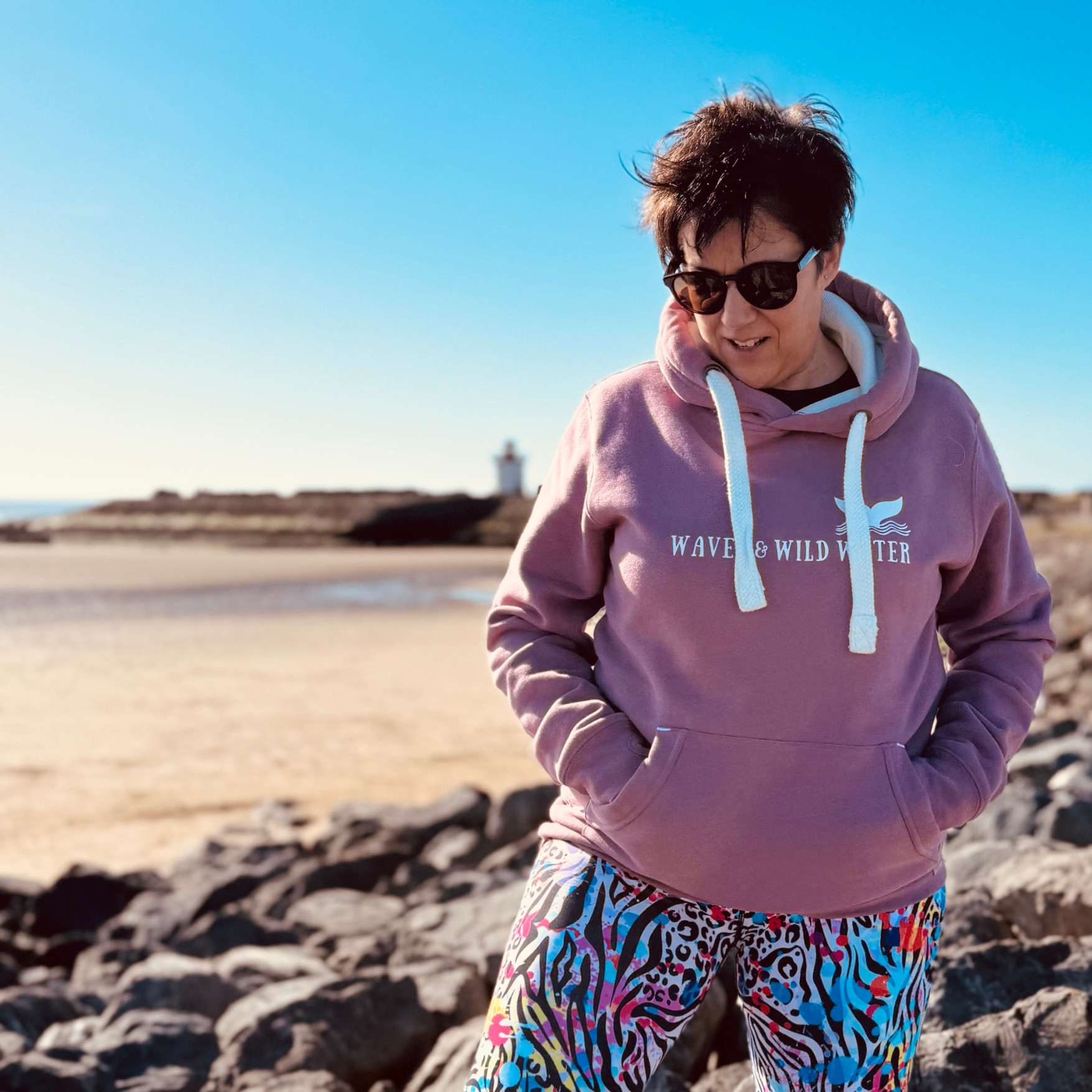 Outdoor swimmer Claire, takes a break from cold sea dipping to explore the rocks. She is wearing her fabulous Waves and Wild Water hoodie in Sunset Blush with white logo print.
