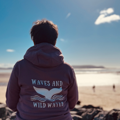Wild swimmer Claire, takes a moment to sit and watch the tides in her Sunset Blush Waves and Wild Water hoodie. The striking whale tail logo is proudly displayed across her back in vibrant white print.