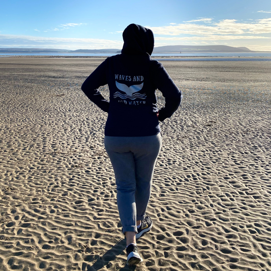 Waves and Wild Water Owner Claire, takes a moment to enjoy the calm beach, which stretches out for miles before her. Keeping the cold at bay, she is snuggled in her navy blue heavyweight hoodie with the whale tail logo on the back in silver ink.