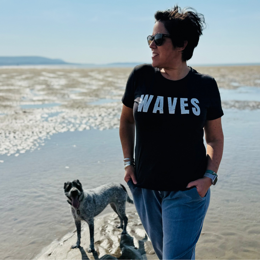 Wild swimmer and owner of Waves and Wild Water, Claire, wears her Just Waves t-shirt in black while being photobombed on the beach by her dog!