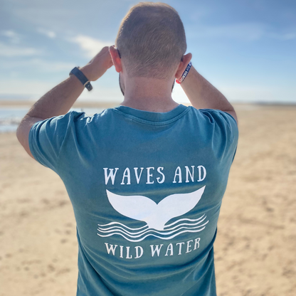 Glenn wears his Seaweed green Waves and Wild Water on a deserted beach after a sunny dip in the sea. The sun picks out the bright white logo across the back, emphasising the magnificent whale tail.