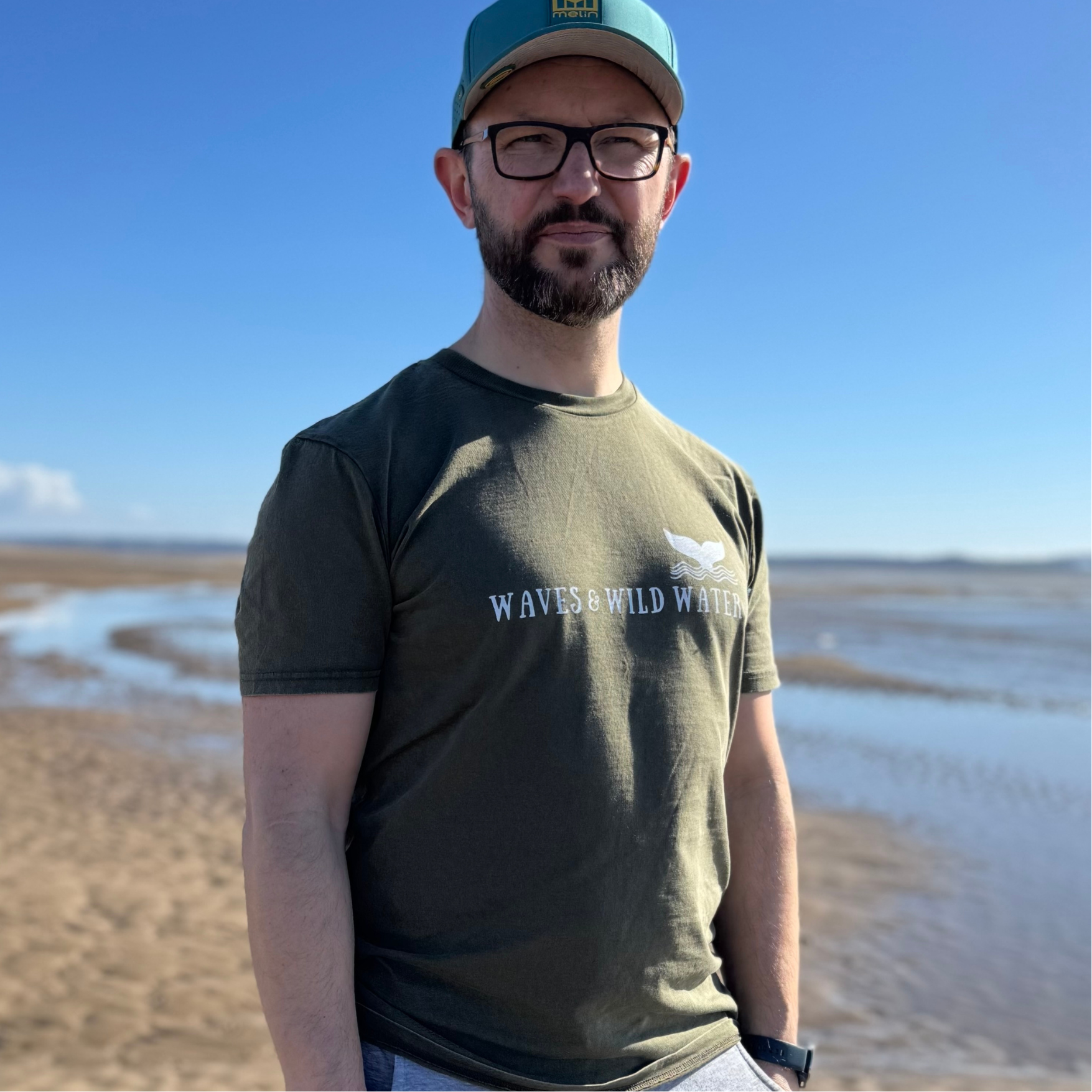 Looking out to sea, wild swimmer Glenn, enjoys a walk on the beach on a bright sunny day. He's wearing his Waves and Wild Water t-shirt in River Bed green, which compliments the bright white logo print beautifully.