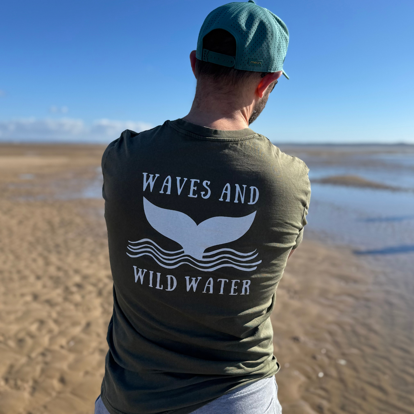 The back of Glenn's waves and Wild Water t-shirt in River Bed green. The bright white whale tail logo print looks great across his back. This tee is LOVED by wild swimmers, outdoor adventurers, and cold water enthusiasts!