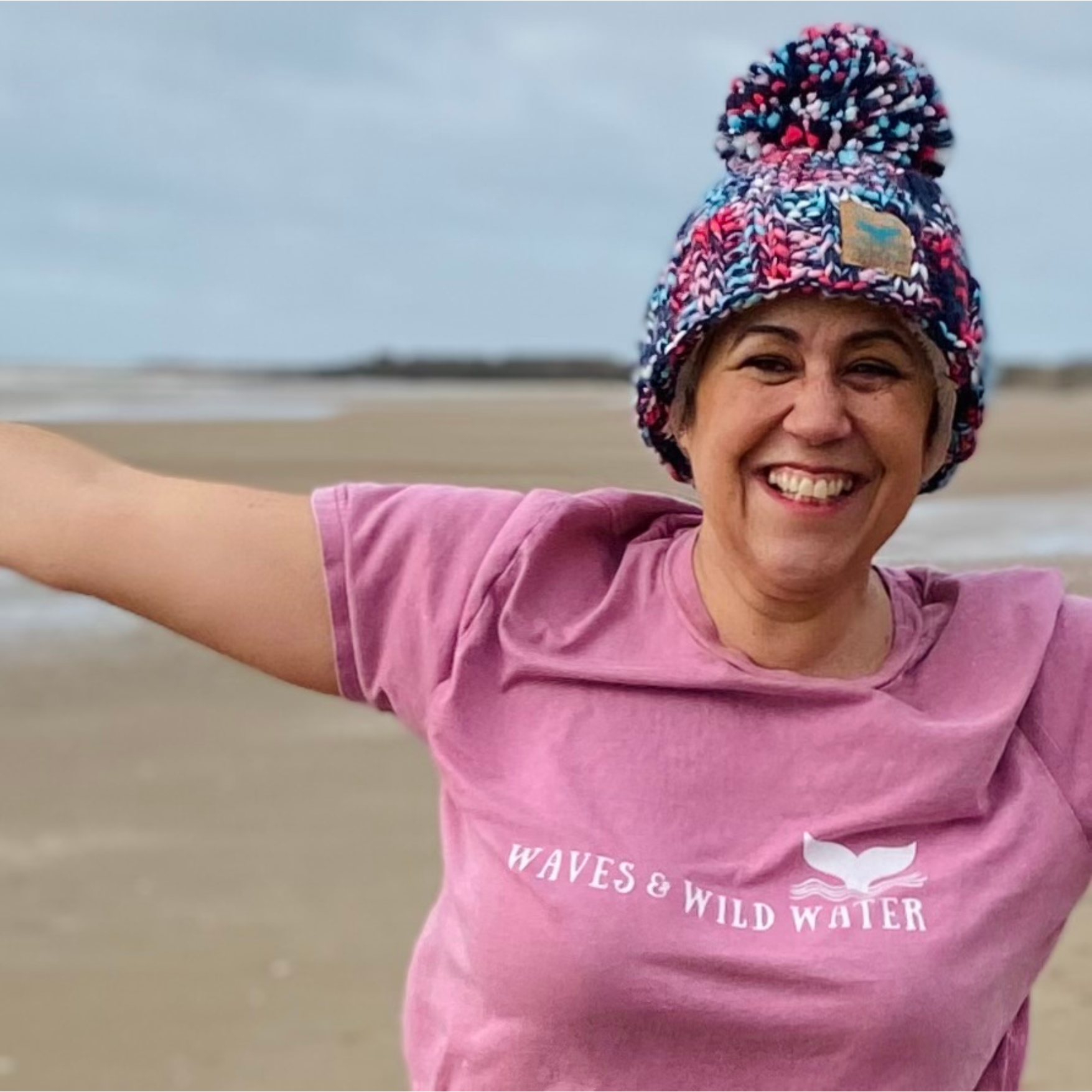 Wild swimmer Claire trying not to get blown away on a cold windy Welsh beach, whilst proudly showing off her Seashell Pink t-shirt.