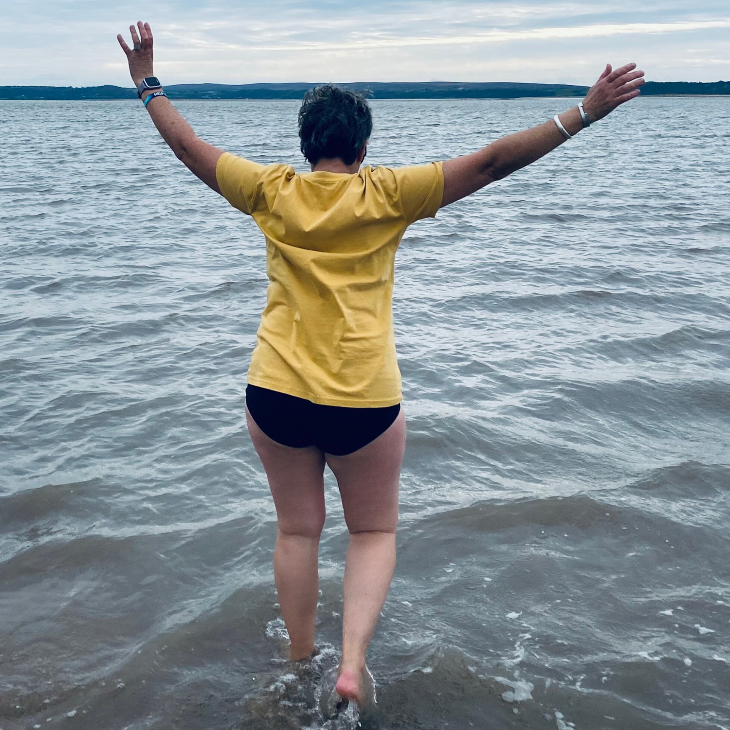 Wild swimmer Claire, wades into the cold sea with her hands in the air, in celebration of seaside rock!