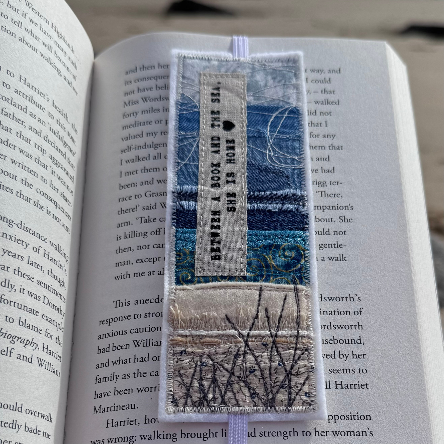 Handmade fabric bookmark resting inside an open book, featuring layered coastal textiles in blues and sand tones with stitched grasses and a printed sea quote — a thoughtful gift for wild swimmers and ocean lovers.