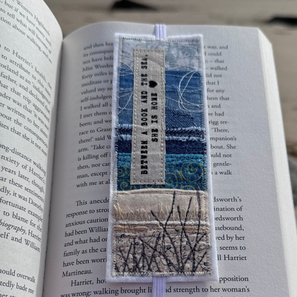 Handmade fabric bookmark resting inside an open book, featuring layered coastal textiles in blues and sand tones with stitched grasses and a printed sea quote — a thoughtful gift for wild swimmers and ocean lovers.