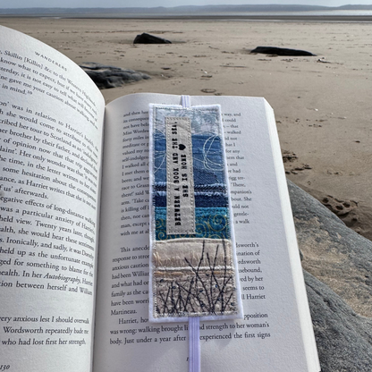 Fabric bookmark placed inside an open book on a sandy beach, showing stitched shoreline design with blue and neutral fabric layers, elastic band, and coastal landscape in the background.