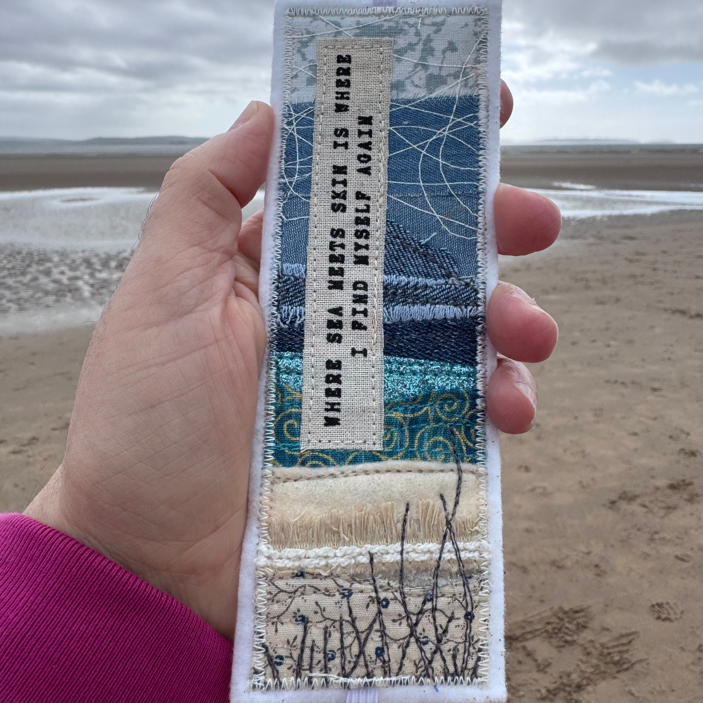 Hand holding a handmade coastal fabric bookmark on a quiet beach, showing stitched shoreline textures and printed quote - a meaningful wild swimming inspired gift made from reclaimed fabrics. Only from Waves & Wild Water.