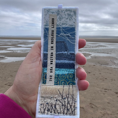 Handmade bookmark held against a tidal beach backdrop, with layered blue fabrics and stitched coastal design — a unique ocean-inspired gift for sea swimmers, readers, and coastal dreamers.