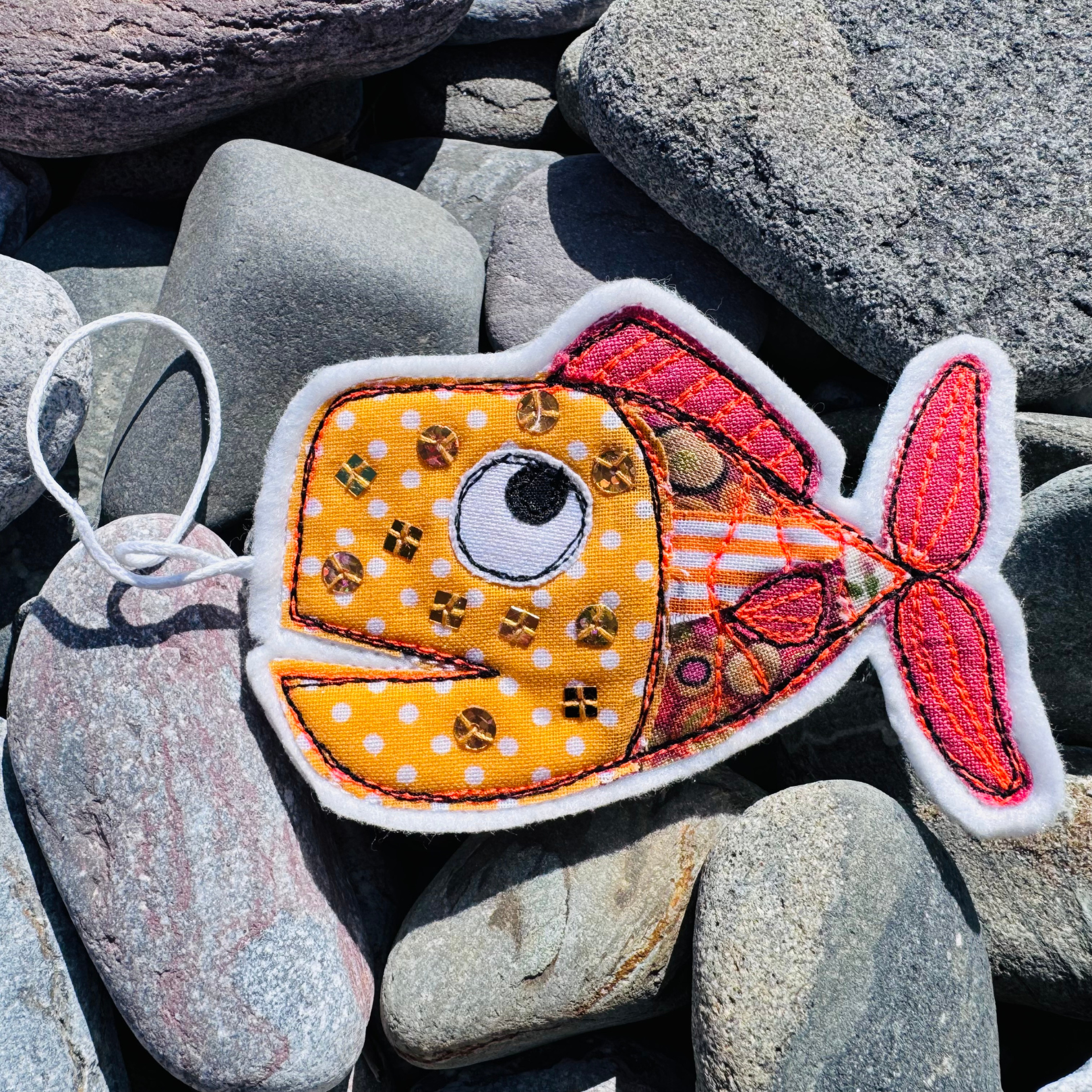 Handmade Fish Wish hanger in orange based fabrics with gold sequins, photographed on smooth coastal stones. Part of the Waves & Wild Water Worthy Waste collection.