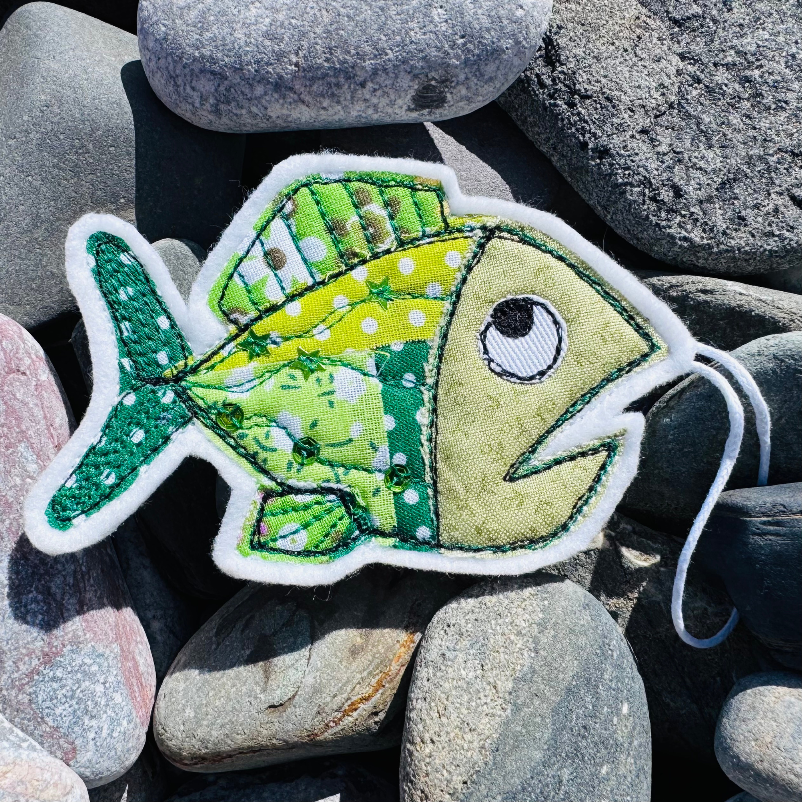 Hand-stitched green Fish Wish hanger made from rescued fabrics with sequin embellishments, displayed on seaside pebbles. Handmade coastal keepsake from Waves & Wild Water.