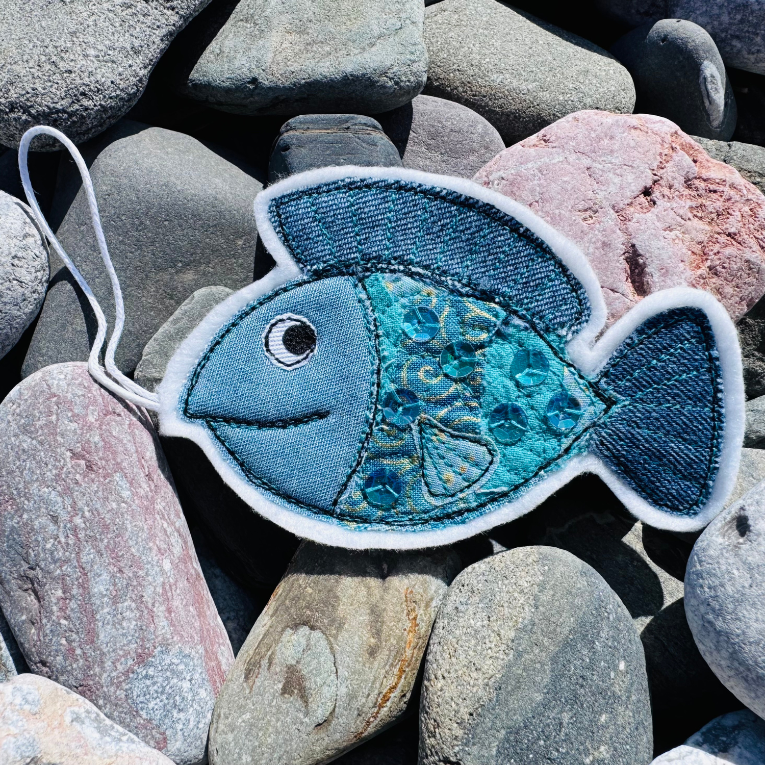Blue based handmade Fish Wish hanger created from upcycled fabrics with sparkling sequins, resting on beach stones. A unique coastal charm from the Waves & Wild Water Worthy Waste collection.