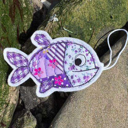Fabric fish ornament in purple tones with patchwork patterns and stitched details, photographed on natural coastal rocks. Handmade in coastal Wales by Waves & Wild Water.
