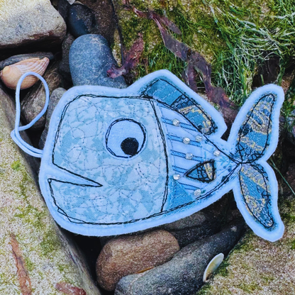 Fish Wish decoration in soft ocean blues and greys, resting among seaweed and stones, a calming coastal keepsake inspired by wild waters.