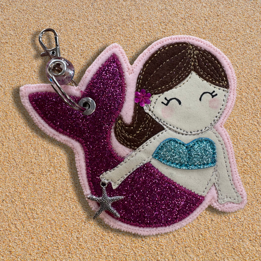 Handmade fabric mermaid charm named Meryl the Mermaid resting on soft golden sand, featuring a glittery pink tail, blue top, and brown hair with flower detail. Key chain clasp has a Murano glass bead. Perfect for hanging from your wild swimming kit bag or changing robe.