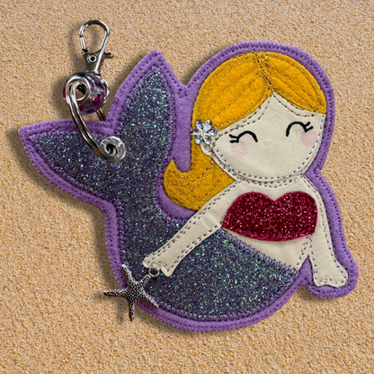 Handmade fabric mermaid charm named Meryl the Mermaid resting on soft golden sand, featuring a glittery lilac tail, pink top, and yellow hair with flower detail. Key chain clasp has a Murano glass bead. Perfect for hanging from your wild swimming kit bag or changing robe.
