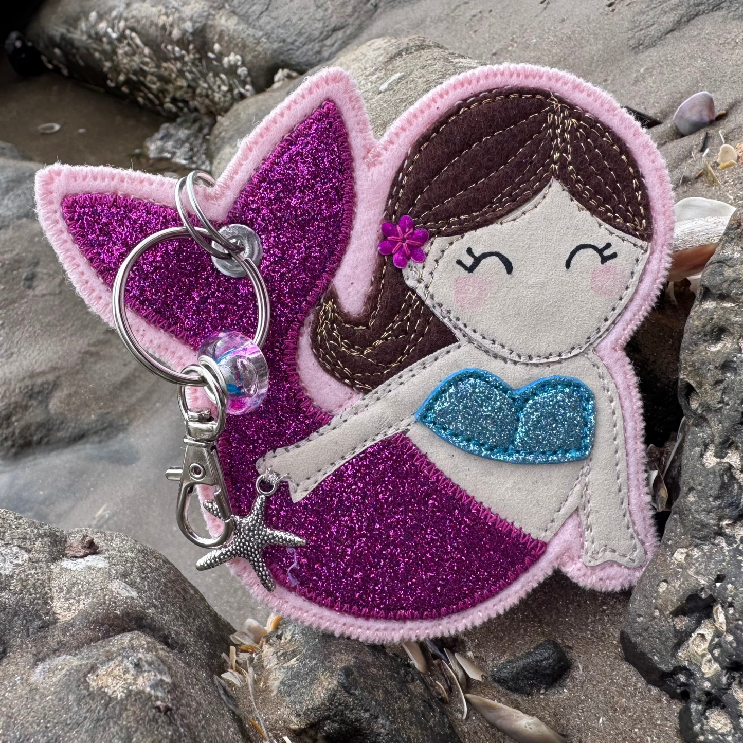 Meryl the Mermaid keyring resting among seaside rocks, her shimmering pink tail catching the light like a warm coastal sunset. Handmade on the coast in Wales for any wild at heart mermaid.