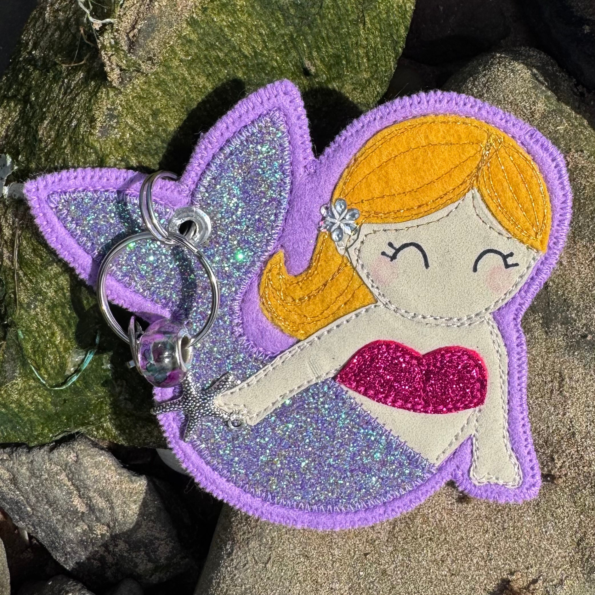 Meryl the Mermaid keyring glowing in soft purples and pinks, resting on sunlit rocks like a little treasure from the tide. Lovingly handmade by the coast in South Wales by Waves & Wild Water.