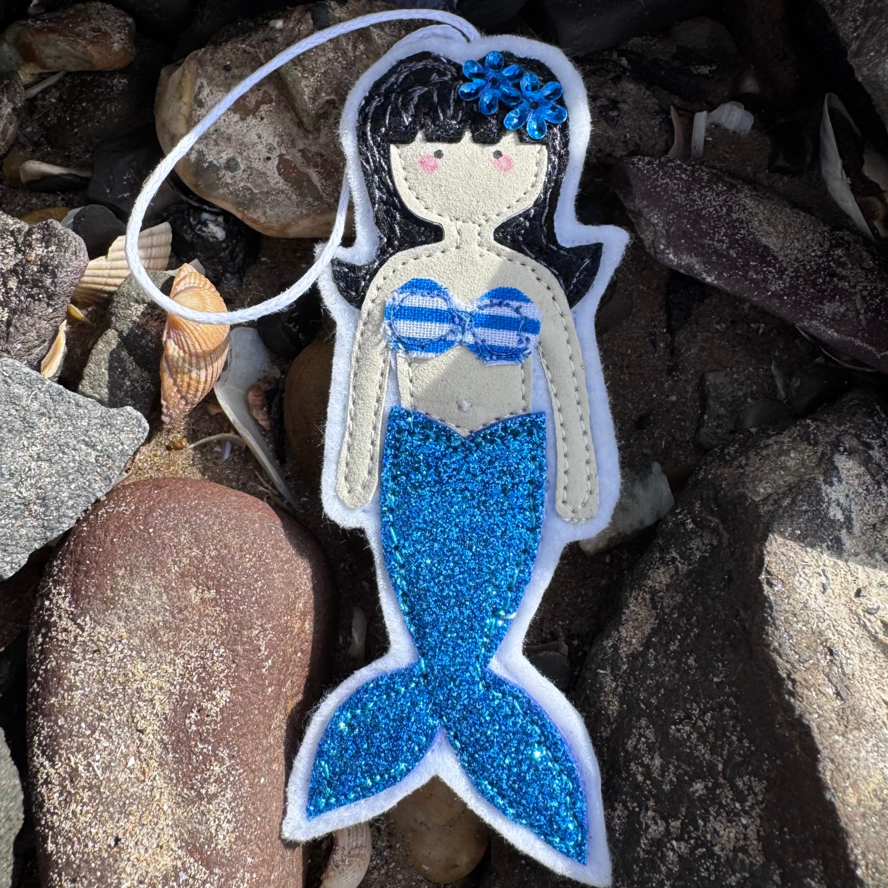 Wild Water Mermaid decoration with bright blue glitter tail resting among shells and seaside stones, a unique handmade gift, created on the coast in Wales.